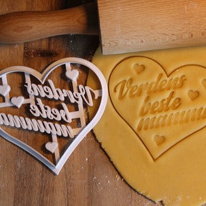 May include: A white plastic heart-shaped cookie cutter with the text "Verdens beste mamma" inside. The cutter is on a wooden surface next to a yellow cookie dough with the same text imprinted on it.