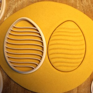 May include: A white plastic cookie cutter in the shape of an egg with a wavy line pattern. The cutter is on top of a yellow cookie dough, which has been pressed with the cutter to create an impression of the wavy line pattern.