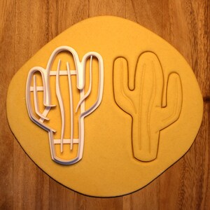 May include: Two white plastic cactus cookie cutters on a yellow surface. The cutters are different sizes and shapes.