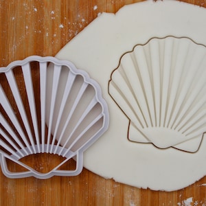 May include: A white plastic seashell cookie cutter on a wooden surface with a white fondant base. The cutter is shaped like a seashell with a ribbed design.