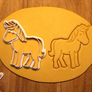May include: Two white cookie cutters in the shape of a unicorn on a yellow surface. The cookie cutters are on top of a rolled out piece of dough.