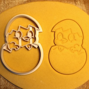 May include: Two cookie cutters, one of a chick hatching from an egg and the other of a chick inside an egg. The cutters are made of white plastic and are on a yellow surface.