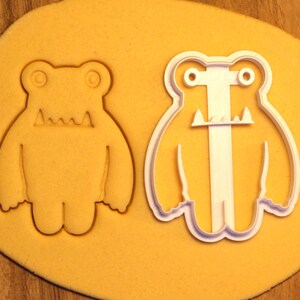 May include: A white plastic cookie cutter in the shape of a monster with two eyes, a mouth with teeth, and arms and legs. The cookie cutter is on a yellow surface.