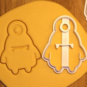 May include: Two white plastic cookie cutters shaped like cartoon monsters. The cutters are on a yellow surface. The cutter on the left has one eye and a mouth with two fangs. The cutter on the right has one eye and a mouth with four fangs.