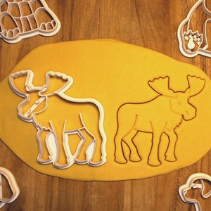 May include: Two white plastic moose cookie cutters on a yellow surface. The cutters are in the shape of a moose with antlers.