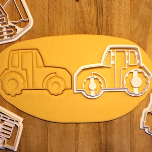 May include: Two white plastic cookie cutters shaped like tractors on a yellow surface. The tractors have large wheels and a detailed design.