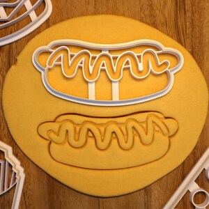 May include: White plastic cookie cutters shaped like hot dogs with mustard. The cutters are on a yellow surface.