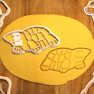 May include: Two white plastic cookie cutters shaped like turtles. The cutters are on a yellow surface.