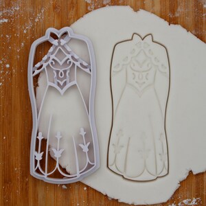 Può includere: Tagliapasta in plastica bianca a forma di vestito con un corpetto dettagliato e accenti floreali. Il tagliapasta è mostrato su una superficie di fondente bianco.