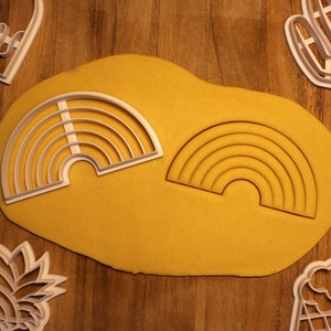 May include: Two white plastic rainbow cookie cutters on a yellow surface. The cookie cutters are on top of a sheet of yellow cookie dough.