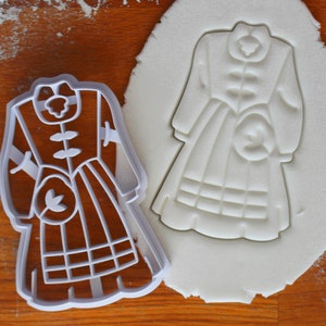 Puede incluir: Cortador de galletas de plástico blanco en forma de vestido con cuello alto, mangas largas y falda a cuadros. El cortador está sobre una superficie blanca.