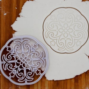 Puede incluir: Cortador de galletas de plástico blanco con un diseño floral detallado. El cortador está sobre una hoja de fondant blanco.