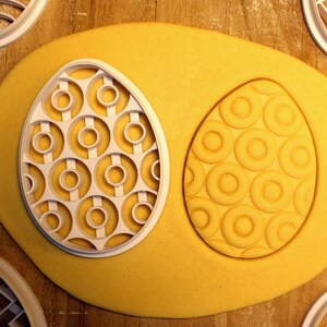 May include: A white plastic cookie cutter shaped like an egg with a circular pattern. The cutter is on top of a yellow dough that has been pressed with the cutter to create an embossed egg shape.