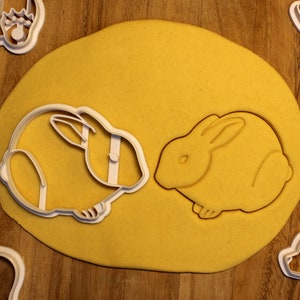 May include: Two white plastic cookie cutters shaped like rabbits on a yellow surface. The cutters are for making cookies.