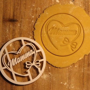 May include: A wooden cookie cutter in the shape of a heart with the word "Mamma" inside. The cutter is on a wooden surface next to a piece of dough with the same design imprinted on it.