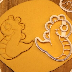 May include: Two white plastic cookie cutters shaped like cartoon seahorses. The seahorses have large eyes, a fin on their backs, and a tail with stripes. The cutters are on a yellow surface.