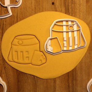 May include: Cookie cutters in the shape of a barrel and cheese, pressed into yellow dough. The barrel cutter has vertical lines and a circular detail. A wedge of cheese is next to the barrel. The cutters are white.