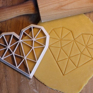 May include: A white plastic heart-shaped cookie cutter with a geometric design. The cutter is placed on top of a sheet of yellow cookie dough with the same geometric design pressed into it.