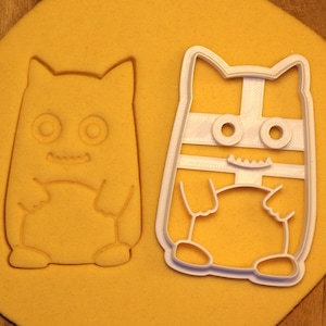 May include: Two white plastic cookie cutters shaped like cartoon monsters. The cutters are on a yellow surface. The monster on the left has a simple design with two eyes, a mouth, and arms. The monster on the right has a more detailed design with a striped body, two eyes, a mouth, and arms.