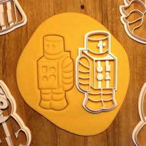 May include: Two white plastic cookie cutters shaped like robots on a yellow surface. The robot on the left has a square, triangle, and circle on its chest. The robot on the right has a rectangular body with a cross on its chest.