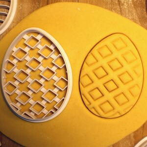 May include: A close-up of a yellow cookie dough with two cookie cutters pressed into it. The cookie cutters are in the shape of a waffle and a grid pattern.