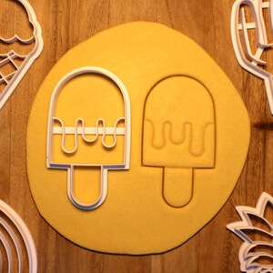 May include: Two white plastic cookie cutters shaped like popsicles with a dripping design on a yellow surface. The cookie cutters are on a yellow surface.