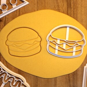 May include: Two white plastic cookie cutters in the shape of a hamburger. The cutters are on a yellow surface.