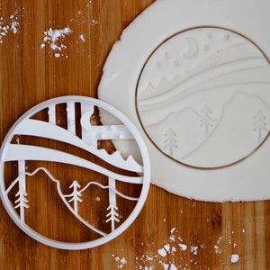 以下が含まれることがあります： 三日月と星が描かれた山脈の形をした白いプラスチック製のクッキーカッター。クッキーカッターは木製の表面の上に置かれています。