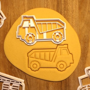 May include: Two white plastic cookie cutters shaped like dump trucks. The cutters are on a yellow surface.