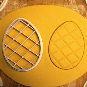 May include: A white plastic cookie cutter in the shape of an egg with a grid pattern, pressed into yellow cookie dough.