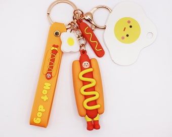 May include: A yellow and orange hot dog keychain with a smiling face, mustard, and a white egg keychain. The keychain also includes a yellow and orange strap with the text "Good Hot Dog" on it.