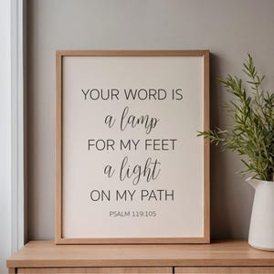 May include: A framed print with the text "YOUR WORD IS a lamp FOR MY FEET a light ON MY PATH PSALM 119:105" in black script. The frame is light brown, and the background is a cream colour. The print is displayed on a wooden surface.