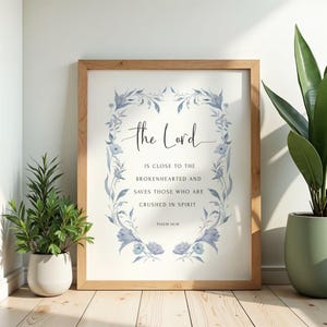 Può includere: Una stampa artistica incorniciata con un bordo floreale ad acquerello azzurro che circonda il testo: The Lord is close to the brokenhearted and saves those who are crushed in spirit. Psalm 34:18 è stampato in piccolo testo nero in basso.