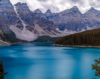 May include: A panoramic view of a mountain lake with snow-capped peaks and a forest of evergreen trees. The water is a vibrant blue and the sky is cloudy.
