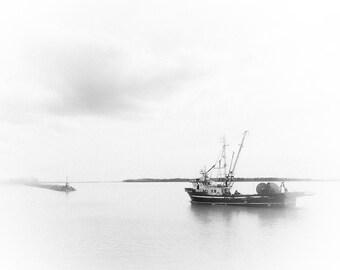 May include: A black and white photo of a fishing boat docked in a harbor. The boat is in the foreground and the water is calm. The sky is cloudy and the horizon is in the distance.