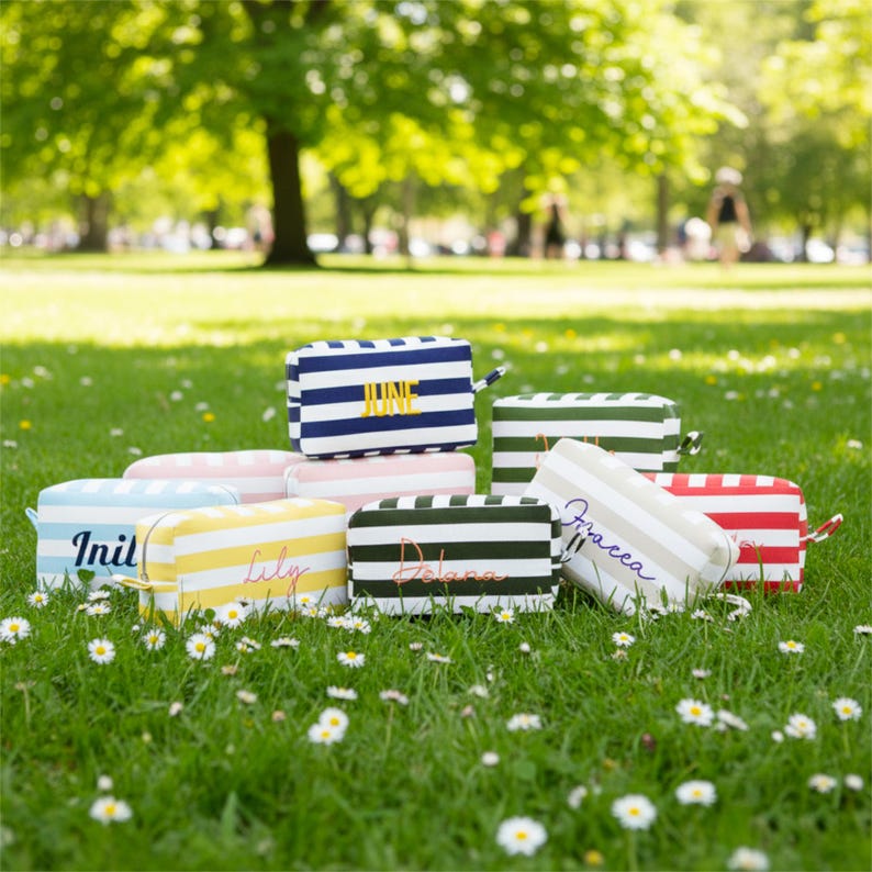 May include: A collection of striped cosmetic bags in various colors, including blue, pink, green, yellow, and red. Each bag features a different name embroidered on the front. The bags are arranged on a bed of green grass with small white flowers.