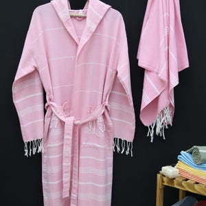 Può includere: Una vestaglia da bagno rosa con cappuccio e strisce bianche e un asciugamano rosa abbinato. Entrambi hanno un bordo con frange bianche.