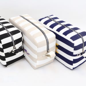 May include: Three rectangular cosmetic bags with horizontal stripes. One is black and white, the second is beige and white, and the third is navy and white. Each bag has a black zipper running across the top.