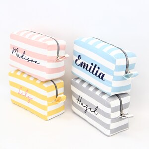 May include: Four rectangular cosmetic bags with horizontal stripes in pink, yellow, blue, and gray. Each bag has a black zipper and a name embroidered on the front: Madison, Lily, Emilia, and Hazel.