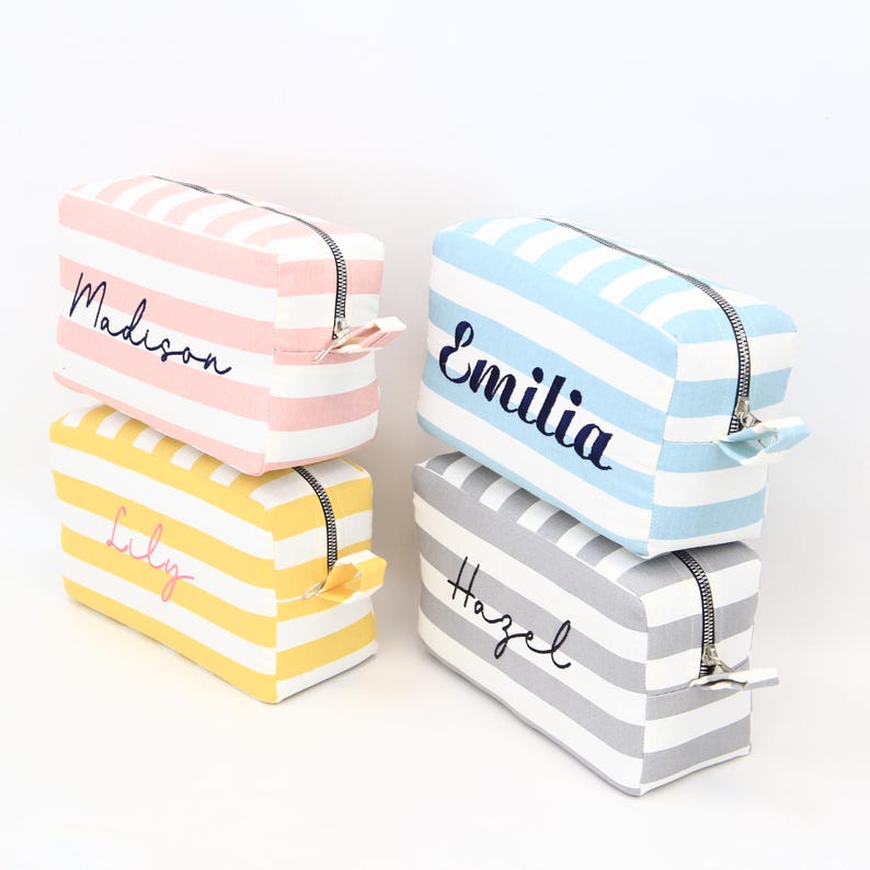 May include: Four rectangular cosmetic bags with horizontal stripes in pink, yellow, blue, and gray. Each bag has a black zipper and a name embroidered on the front: Madison, Lily, Emilia, and Hazel.