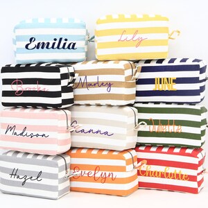 May include: A stack of personalized cosmetic bags with horizontal stripes in various colors, including blue, yellow, black, tan, pink, white, orange, gray, green, and red. Each bag has a name embroidered on it in a cursive font.