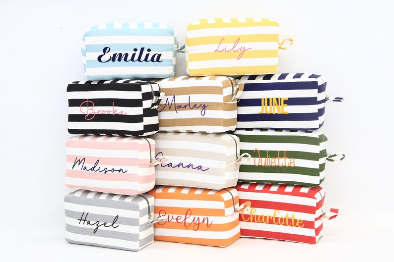 May include: A stack of personalized cosmetic bags with horizontal stripes in various colors, including blue, yellow, black, tan, pink, white, orange, gray, green, and red. Each bag has a name embroidered on it in a cursive font.