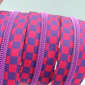 Puede incluir: Primer plano de una cinta de cremallera con un patrón a cuadros en rojo y morado. La cinta de cremallera tiene una bobina de cremallera de metal rosa.