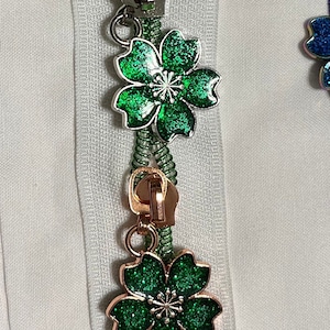 Puede incluir: Un tirador de cremallera verde con un amuleto de flor verde brillante. El tirador de cremallera está unido a una cremallera verde.