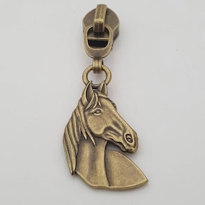 May include: Antique brass zipper pull with a horse head design. The horse head is detailed with a flowing mane and a realistic profile. The zipper pull is attached to a metal slider.