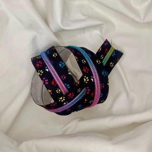 May include: A black zipper with a repeating pattern of colorful paw prints. The zipper teeth are a gradient of rainbow colors, and the zipper tape is folded over itself.