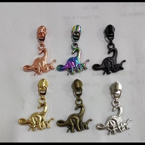 May include: A selection of dinosaur-shaped zip pulls in various metallic finishes. The pulls include rose gold, iridescent, black, gold, bronze, and silver. Each pull features a detailed dinosaur design.