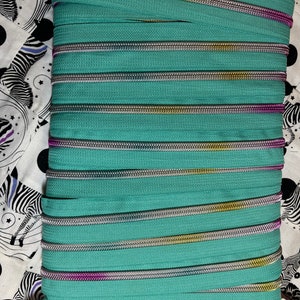 以下が含まれることがあります： ターコイズブルーのジッパーのクローズアップ。シルバー、ゴールド、パープルのアクセントが施されています。ジッパーは複数のストランドで構成されており、白い表面に平らに置かれています。