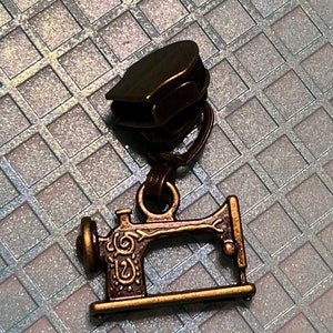 May include: A bronze-colored zipper pull with a decorative sewing machine charm. The zipper pull is a dark brown color, and the charm is a detailed miniature of a vintage sewing machine.