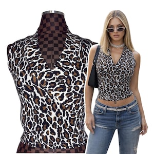 May include: A sleeveless halter top with a V-neckline, featuring a leopard print pattern in brown, black, and white. The top is displayed on a mannequin and worn by a person with sunglasses and blue jeans.